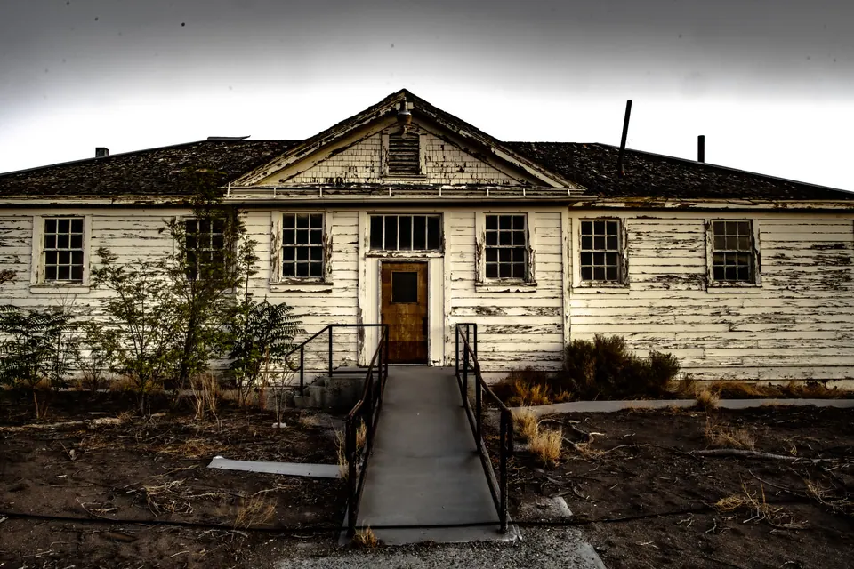 A weathered white building faces an overgrown path beneath a grey sky.