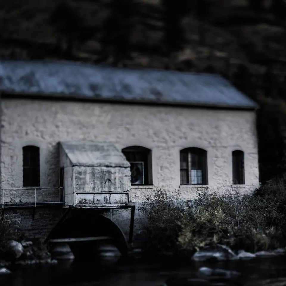 A pale stone building and an old wheel stand beside dark water and trees.