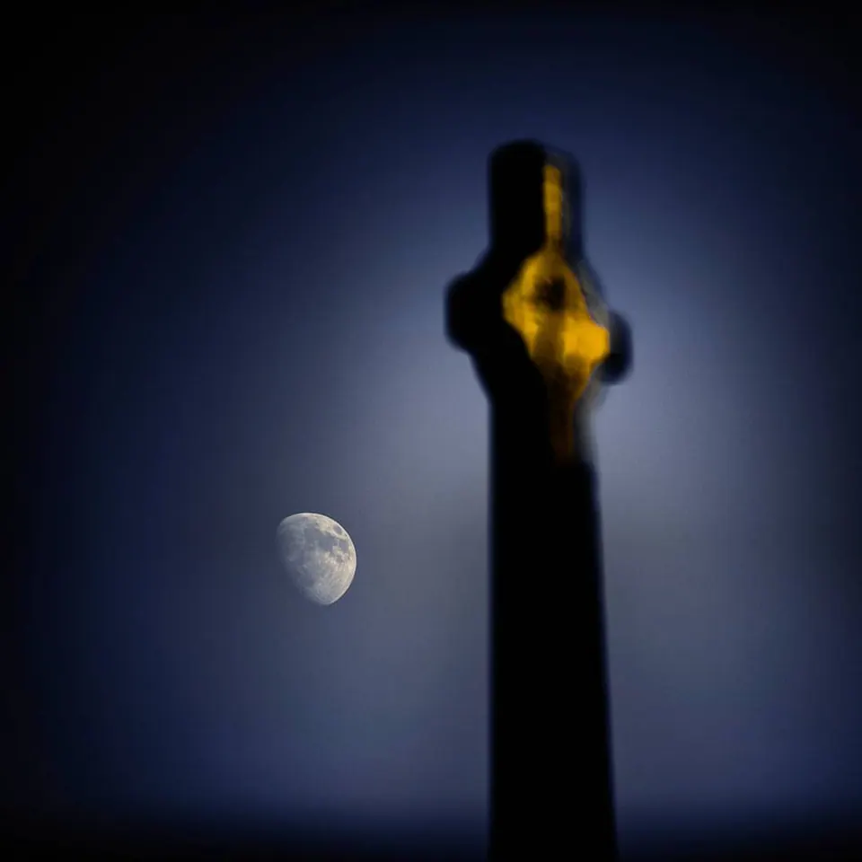 A pale moon hangs in a blue sky beside a softly blurred cross.