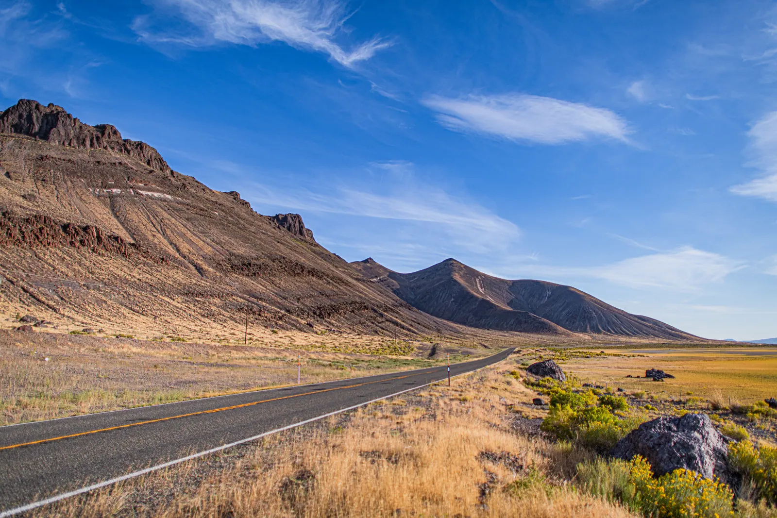 An empty road curves past a broad, rocky ridge beneath a bright sky.