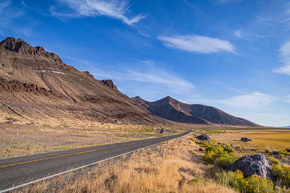 An empty road curves past a broad, rocky ridge beneath a bright sky.