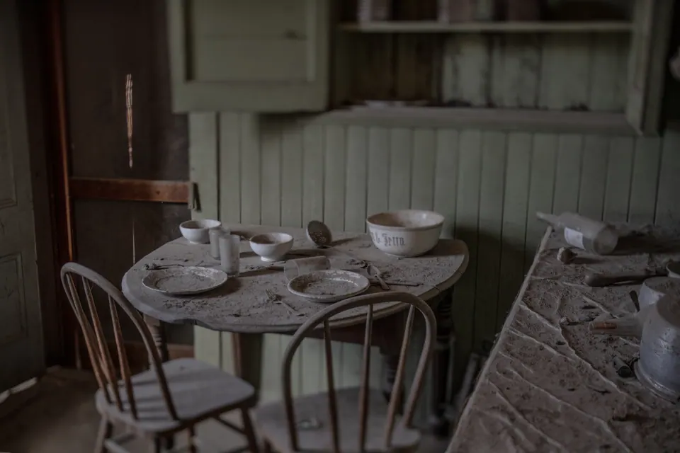 Cups and bowls remain on a small table in an abandoned wooden kitchen.