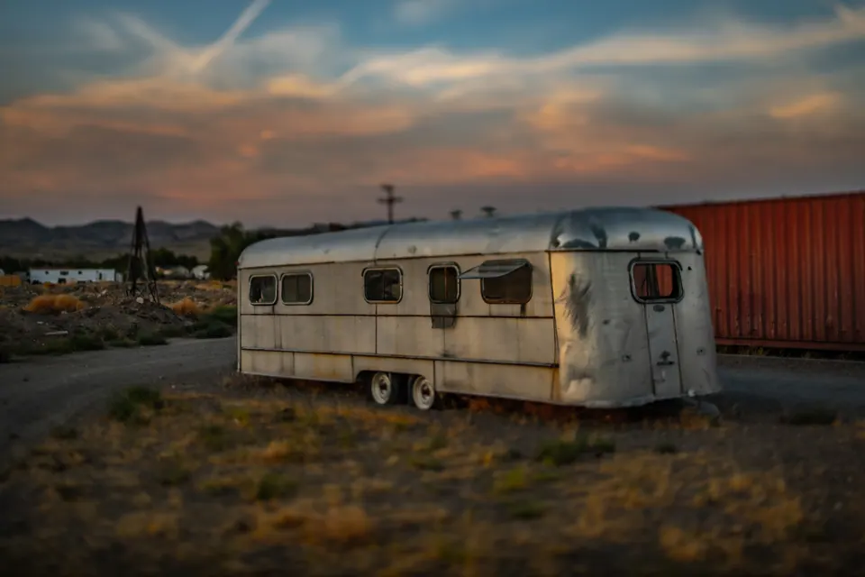 A small weathered trailer rests among dry grass beneath an evening sky.