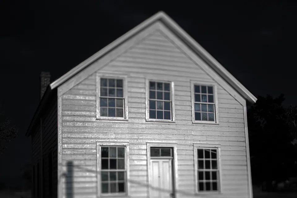 A simple white wooden house stands out against a dark background in black and white.