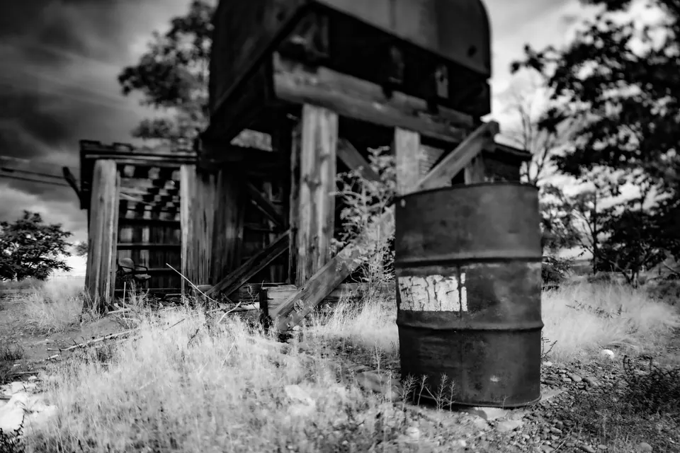 An old wooden structure and a metal barrel stand in overgrown grass, rendered in black and white.