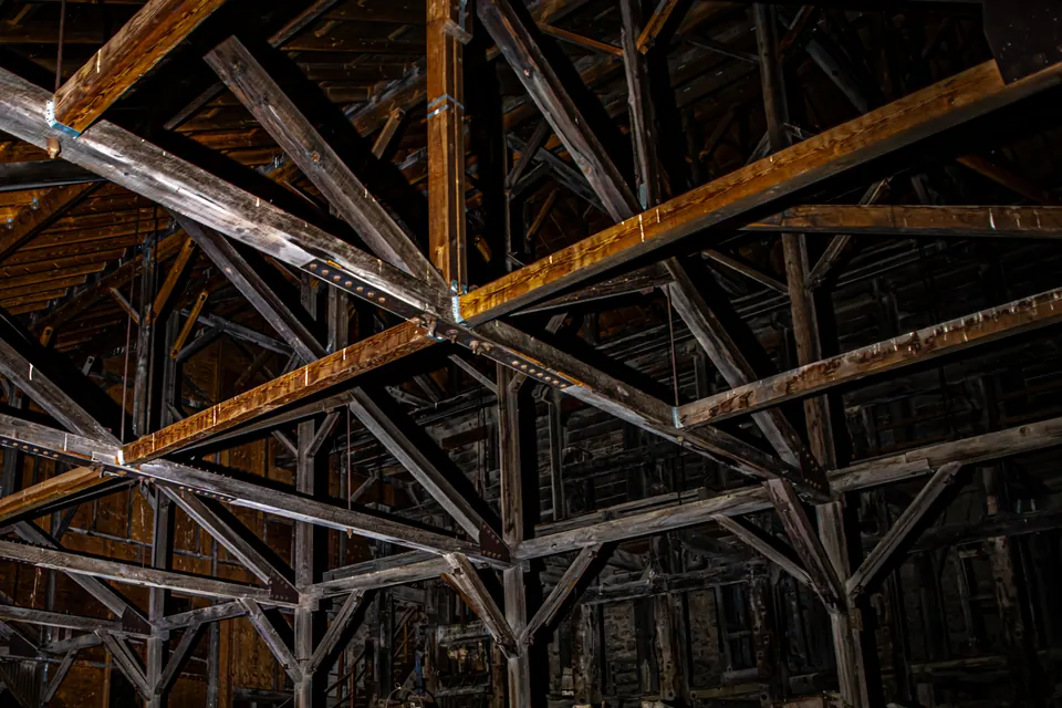 Heavy wooden rafters crisscross the dark interior of a large old building.