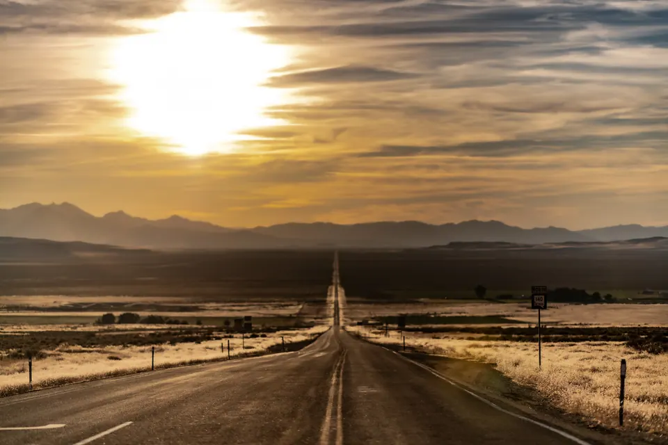 A straight desert road reaches toward distant mountains under a low, golden sun.