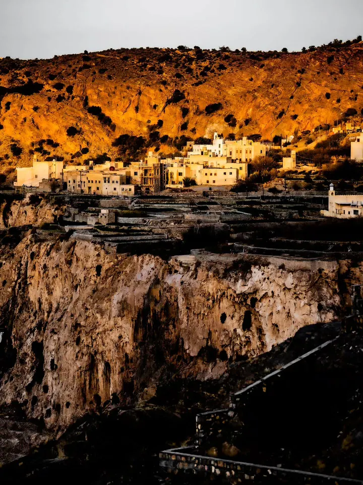 A pale village sits below a glowing ochre hillside as the valley falls into shadow.
