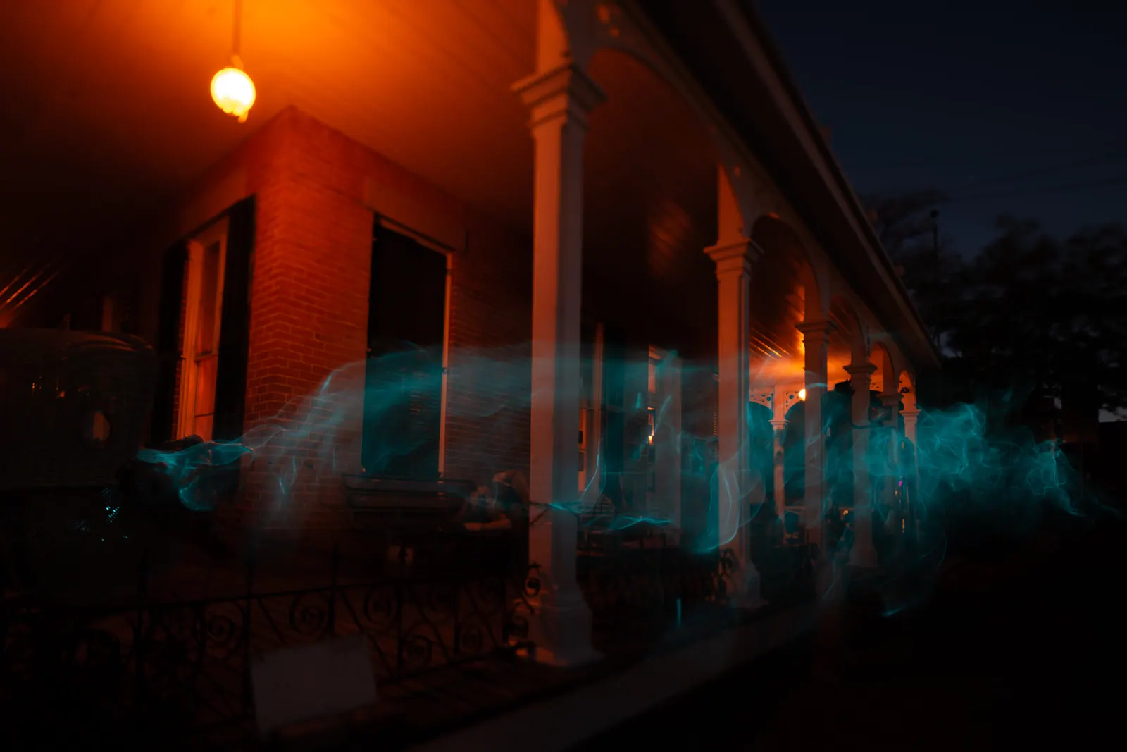 Blue light trails pass along the warmly lit porch of a house at night.