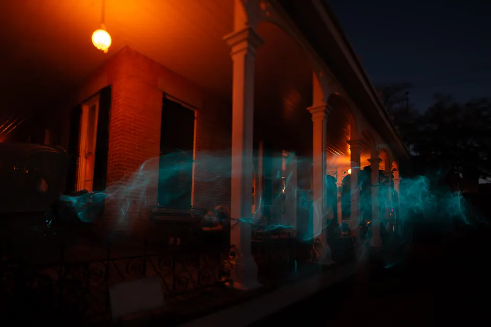 Blue light trails pass along the warmly lit porch of a house at night.