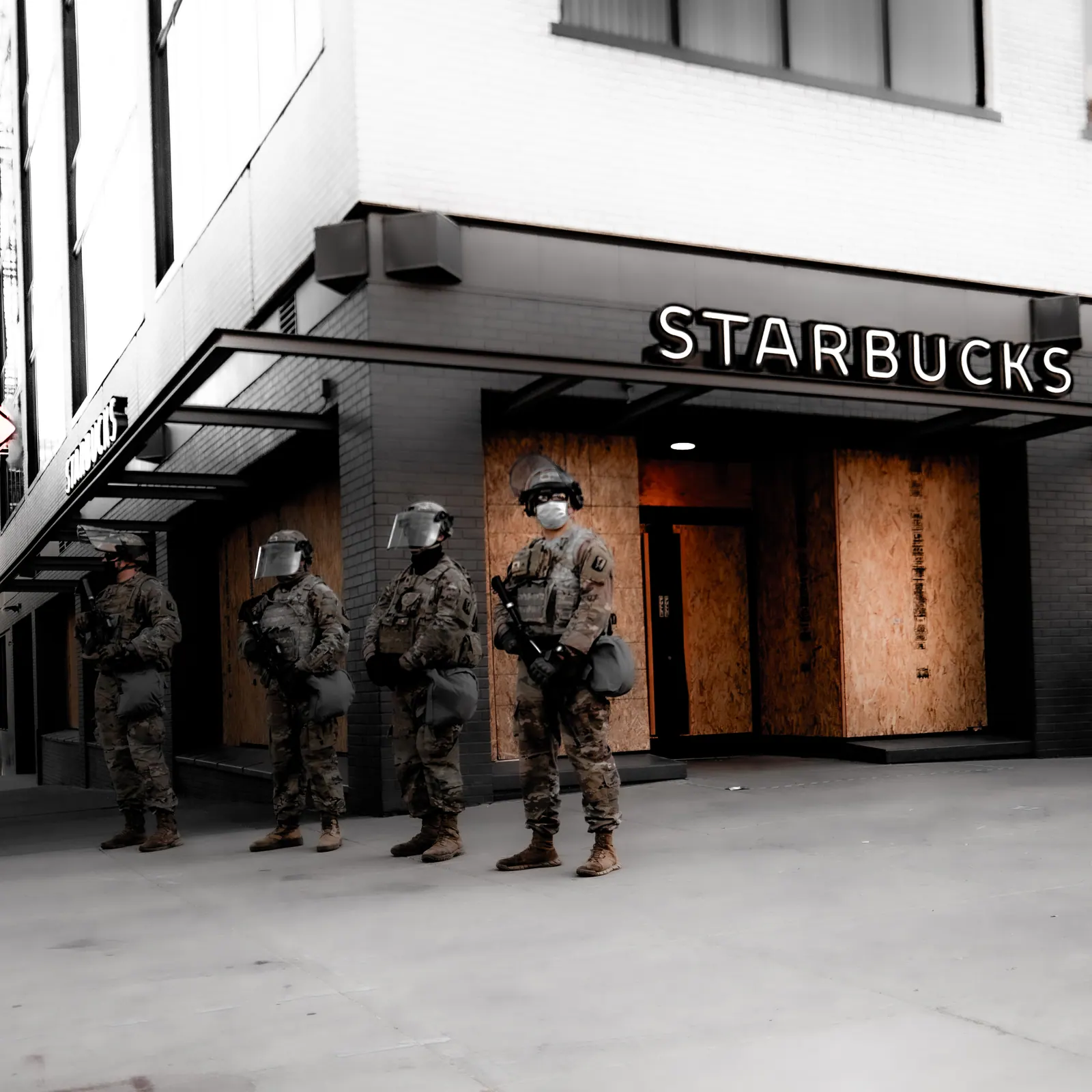 Uniformed figures stand beside a coffee shop on a city street in black and white.