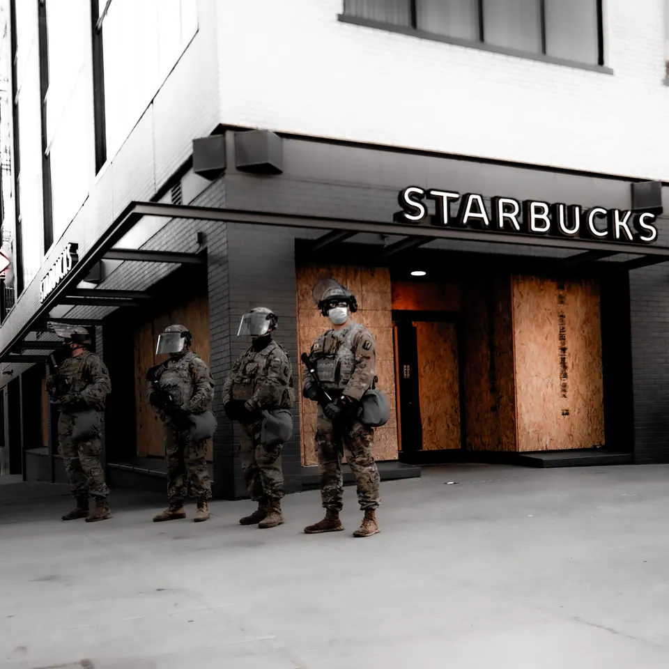Uniformed figures stand beside a coffee shop on a city street in black and white.