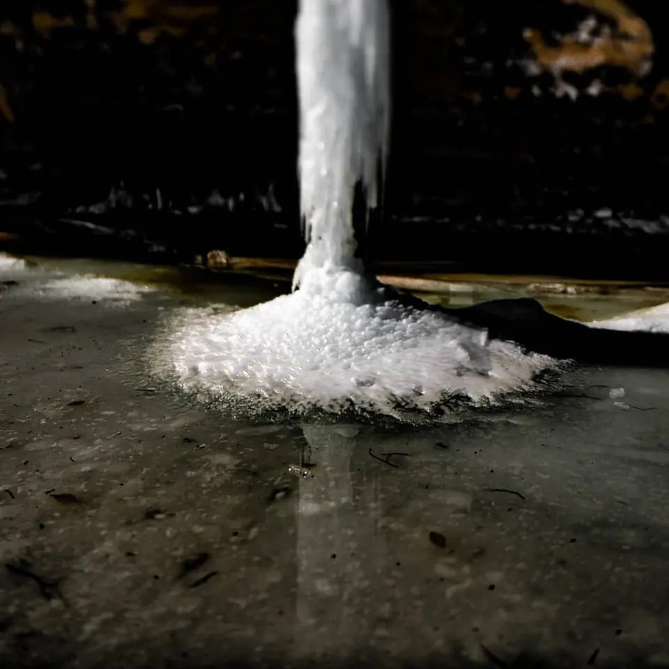 A thick column of ice meets a frozen pool in the shadow of a structure.