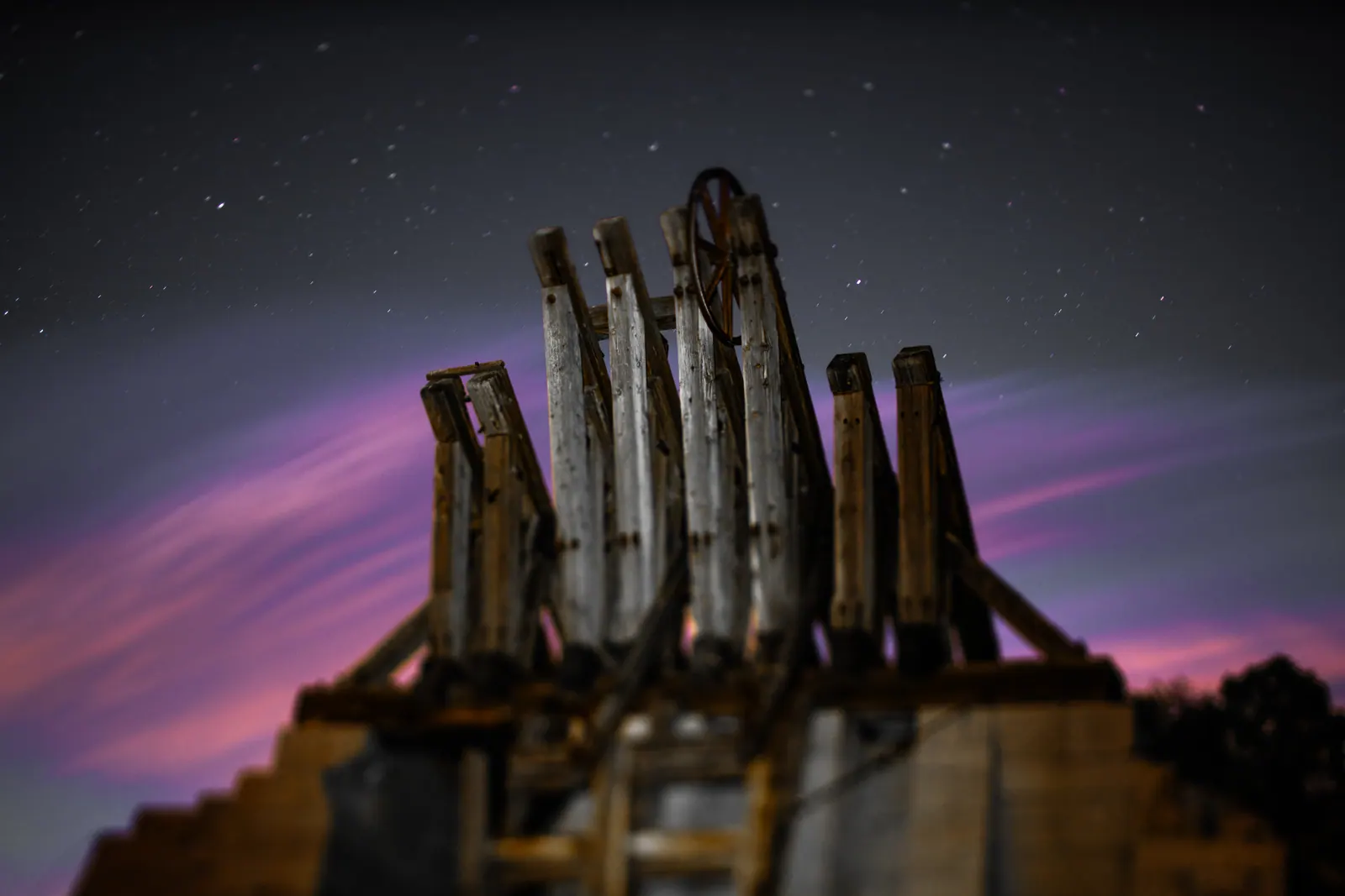Weathered wooden pilings rise into a starry sky streaked with violet and amber light.