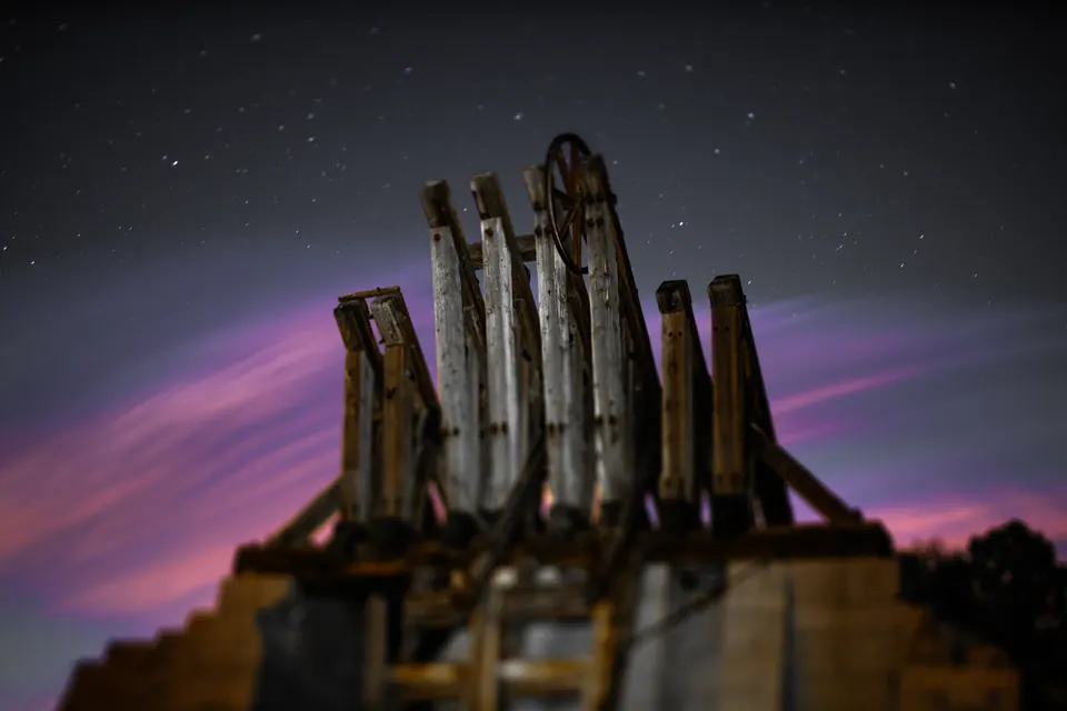 Weathered wooden pilings rise into a starry sky streaked with violet and amber light.