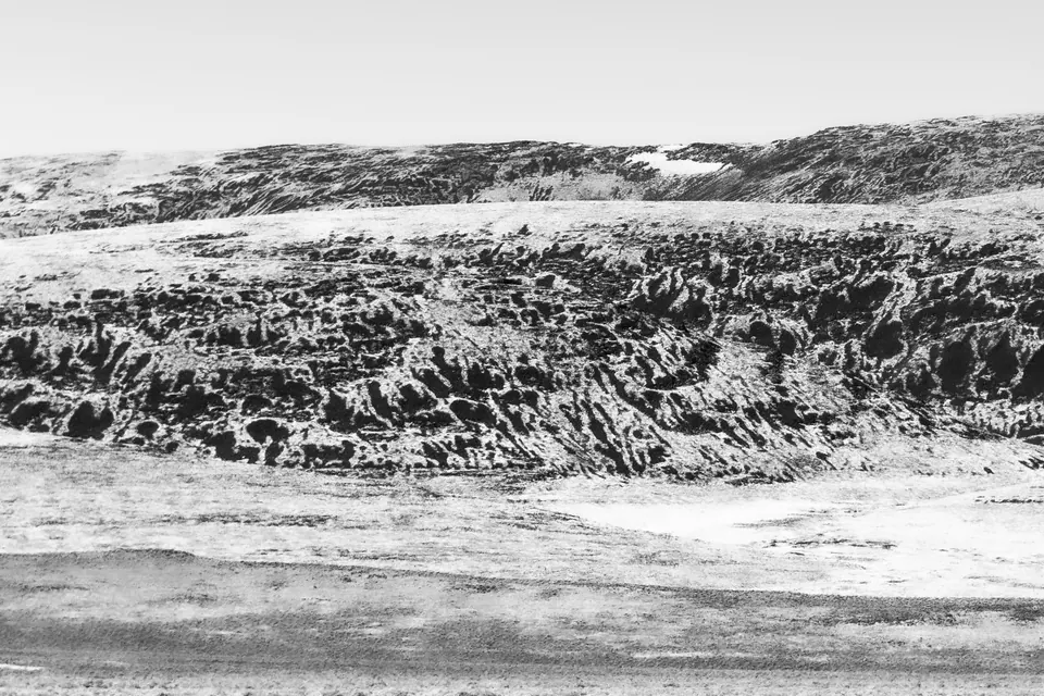Pale light flattens a broad, scrub-covered hillside into a stark monochrome landscape.