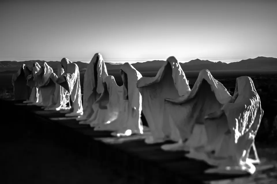 A row of pale, robed sculptures stands in a stark desert landscape.
