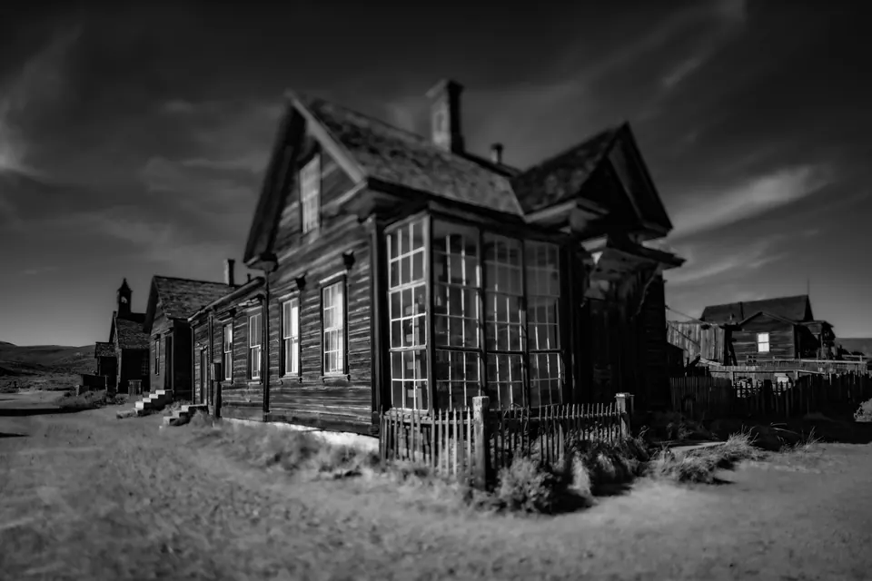 An abandoned wooden building with tall windows stands beneath a dramatic grey sky.