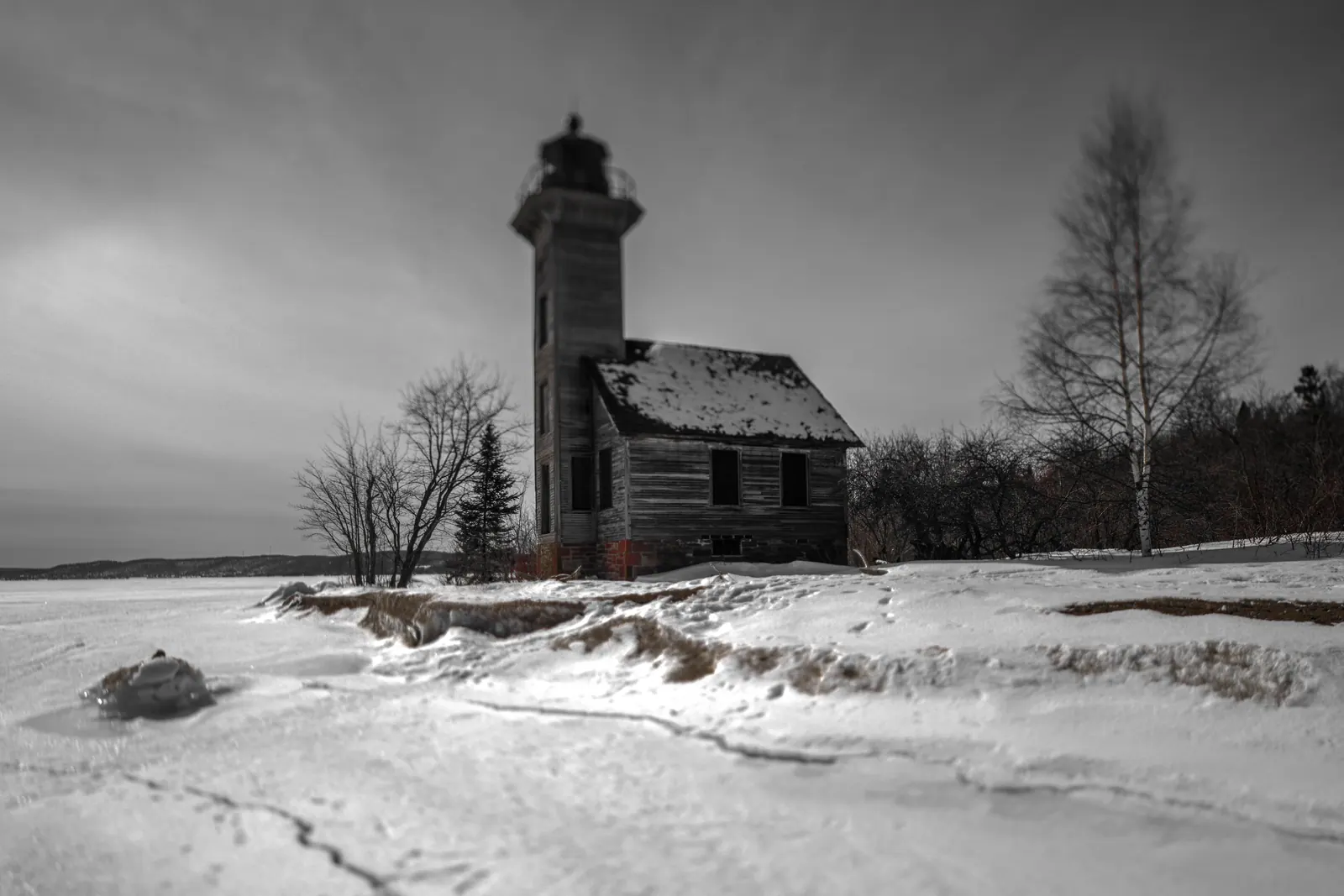 A small lighthouse stands beside a frozen shore in a snowy monochrome landscape.
