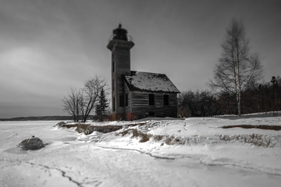 A small lighthouse stands beside a frozen shore in a snowy monochrome landscape.
