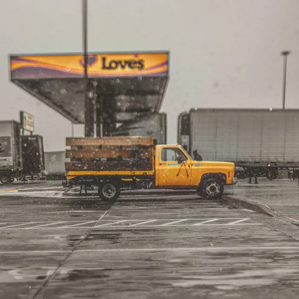 A yellow truck sits below a roadside sign as snow falls across the scene.